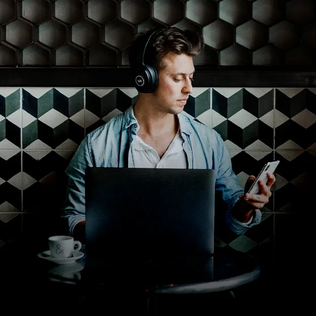 Um homem usando fones de ouvido, sentado a uma mesa pequena com um notebook, olhando o celular.