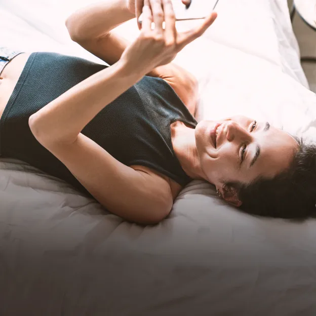 Uma mulher deitada na cama, sorrindo enquanto olha o celular.
